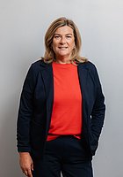 Eine Frau lacht in die Kamera. Sie trägt einen dunkelblauen Hosenanzug und ein rotes Shirt.