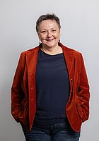 Eine Frau mit Kurzhaarschnitt lacht in die Kamera. Sie trägt ein dunkelblaues Oberteil und einen roten Blazer darüber.