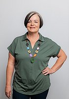 Eine Frau mit Kurzhaarschnitt lacht in die Kamera. Sie trägt eine grüne Bluse und eine bunte Halskette.