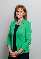 Eine Frau lacht in die Kamera. Sie trägt ein schwarzes Oberteil und einen grünen Blazer.