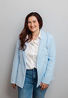 Eine Frau mit langen Haaren lacht in die Kamera. Sie trägt eine weiße Bluse und einen hellblauen Blazer.