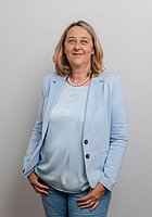 Eine Frau lacht in die Kamera. Sie trägt eine hellblaue Bluse und einen hellblauen Blazer sowie eine Perlenhalskette.