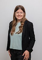 Eine Frau mit langen Locken lacht in die Kamera. Sie trägt ein hellblaues Shirt und einen schwarzen Blazer.