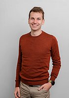 Ein Mann lacht in die Kamera. Er trägt einen roten Pullover und eine beige Hose.