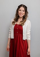 Eine Frau mit langen Locken lacht in die Kamera. Sie trägt ein rotes Kleid und eine weiße Weste darüber.