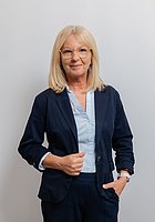 Eine Frau mit Brille lacht in die Kamera. Sie trägt einen dunkelblauen Hosenanzug und eine hellblaue Bluse.