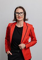 Eine Frau mit Brille lacht in die Kamera. Sie trägt ein schwarzes Oberteil und einen roten Blazer.