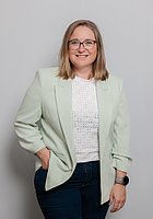 Eine Frau mit Brille lacht in die Kamera. Sie trägt ein weißes Oberteil und einen mintgrünen Blazer.