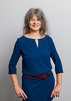 Eine Frau mit gelockten Haaren lacht in die Kamera. Sie trägt ein dunkelblaues Kleid mit einem roten Gürtel.