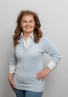 Eine Frau mit langen Locken lacht in die Kamera. Sie trägt einen hellblauen Pullover mit einer weißen Bluse darunter.