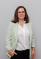 Eine Frau mit Brille lacht in die Kamera. Sie trägt eine weiße Bluse und einen mintgrünen Blazer.