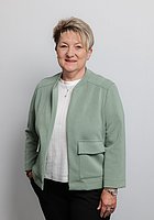 Eine Frau mit Kurzhaarschnitt lacht in die Kamera. Sie trägt ein weißes Oberteil und einen mintgrünen Blazer darüber.