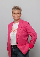 Eine Frau mit Kurzhaarschnitt und Brille lacht in die Kamera. Sie trägt einen pinken Blazer und hält eine Hand in die Hüfte gestützt.