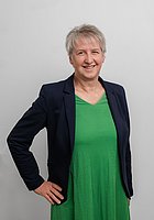 Eine Frau mit Kurzhaarschnitt lacht in die Kamera. Sie trägt ein grünes Kleid und einen schwarzen Blazer.