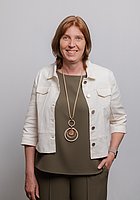 Eine Frau mit einer langen Kette um den Hals lacht in die Kamera. Sie trägt einen olivgrünen Zweiteiler und eine weiße Jacke darüber.