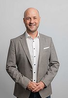 Ein Mann lacht in die Kamera. Er trägt ein weißes Hemd und ein grau-braunes Sakko.