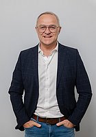 Ein Mann mit Brille lacht in die Kamera. Er trägt ein weißes Hemd mit Sakko, beide Hände stecken in den seitlichen Hosentaschen.