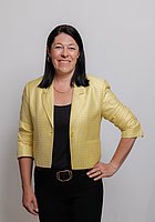 Eine Frau mit schwarzen Haaren lacht in die Kamera. Sie trägt einen gelben Blazer und ein schwarzes Oberteil.