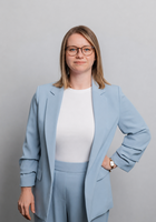 Eine Frau mit Brille lächelt in die Kamera. Sie trägt ein weißes Shirt und einen hellblauen Hosenanzug.
