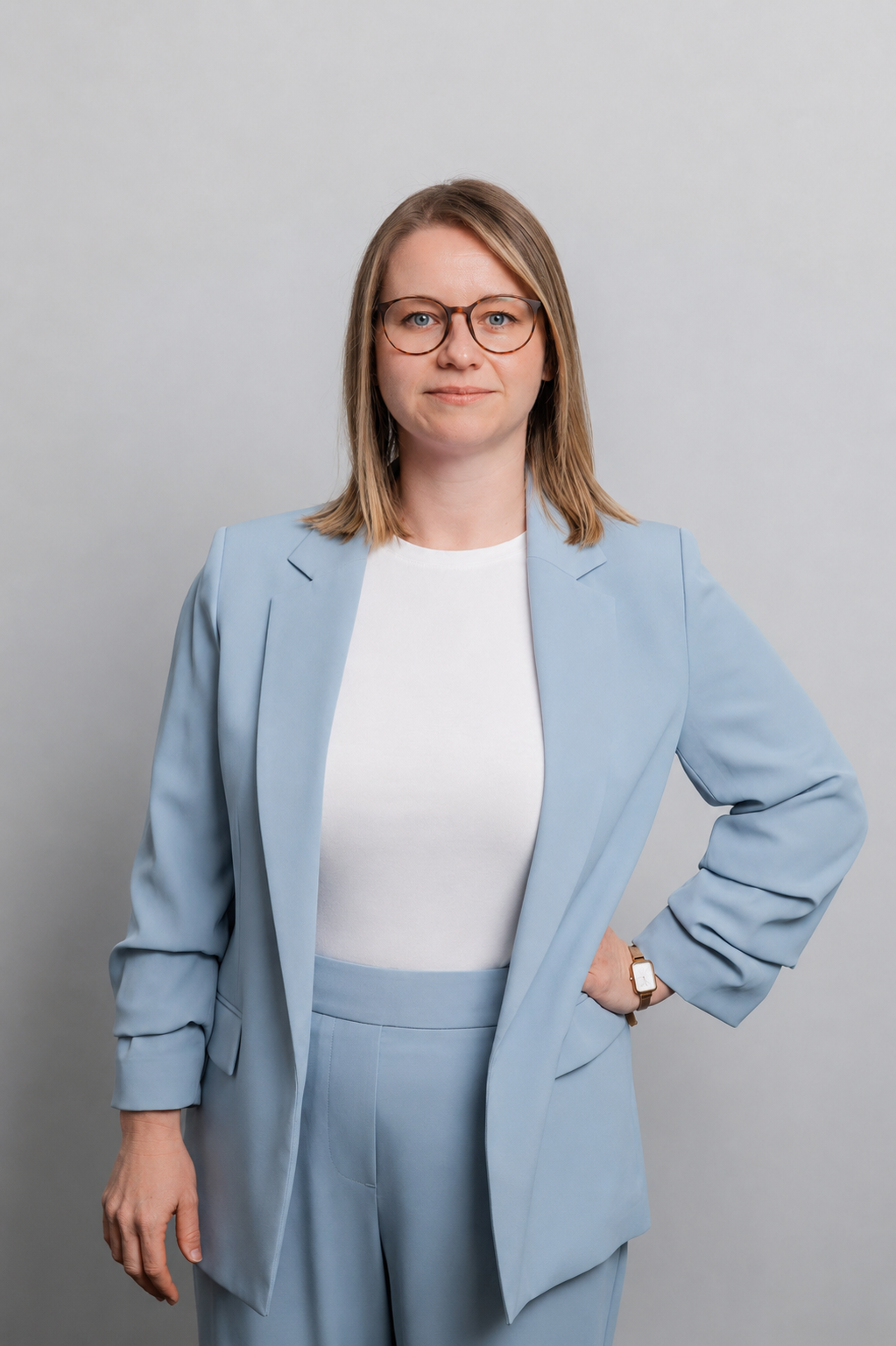 Eine Frau mit Brille lächelt in die Kamera. Sie trägt ein weißes Shirt und einen hellblauen Hosenanzug.