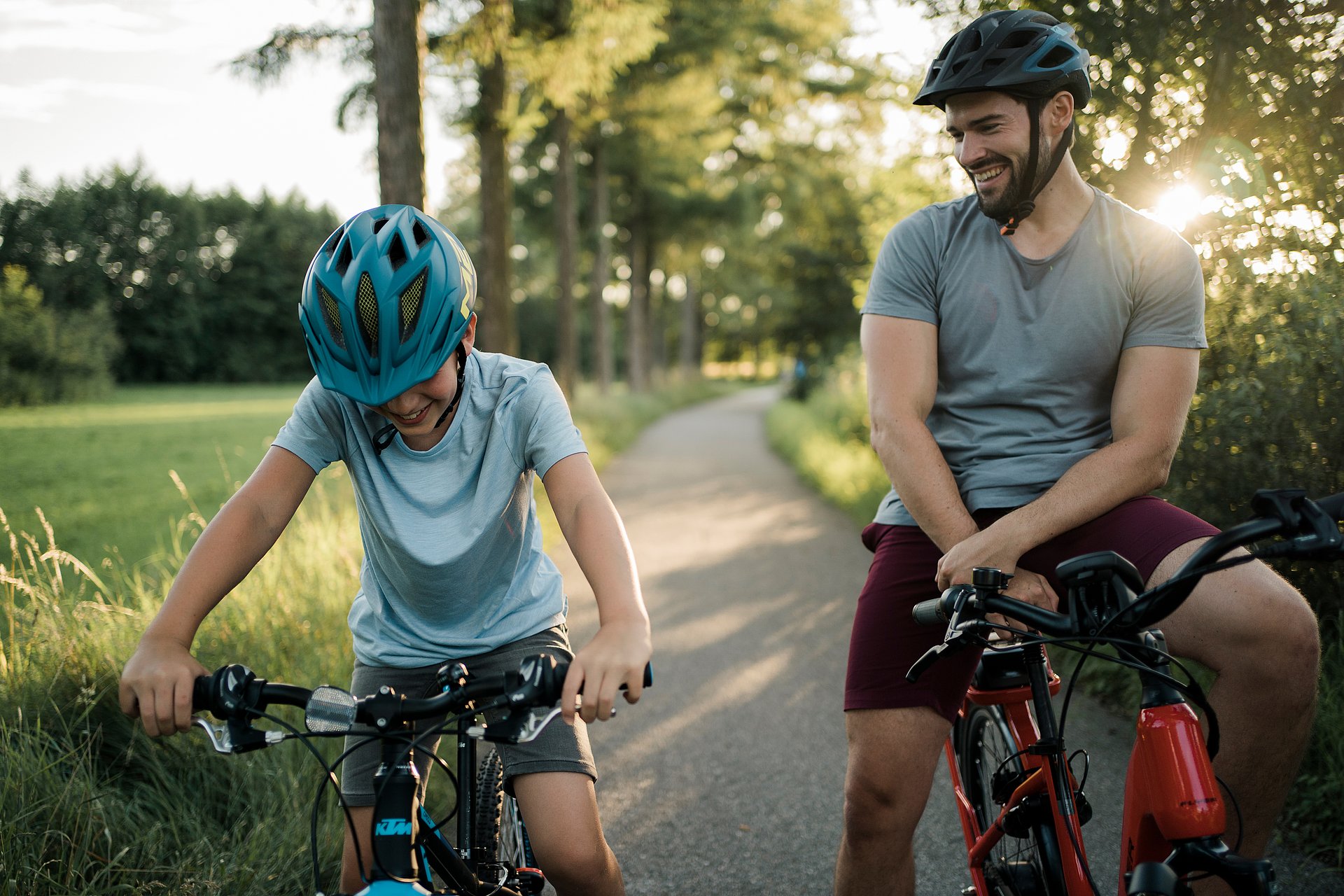 Vater und Sohn sind mit ihren R&auml;dern auf dem Trattnachtal-Radweg unterwegs und machen gerade eine Pause. Sie rasten auf ihren stehenden R&auml;dern, der Vater blickt stolz zu seinem Sohn hin. Dieser zieht an den Handbremsen seines Rades und blickt dorthin. Beide tragen bequeme Bekleidung und Radhelme. Der asphaltierte Radweg ist von Wiesen und B&auml;umen umgeben, im Hintergrund die Sonne.