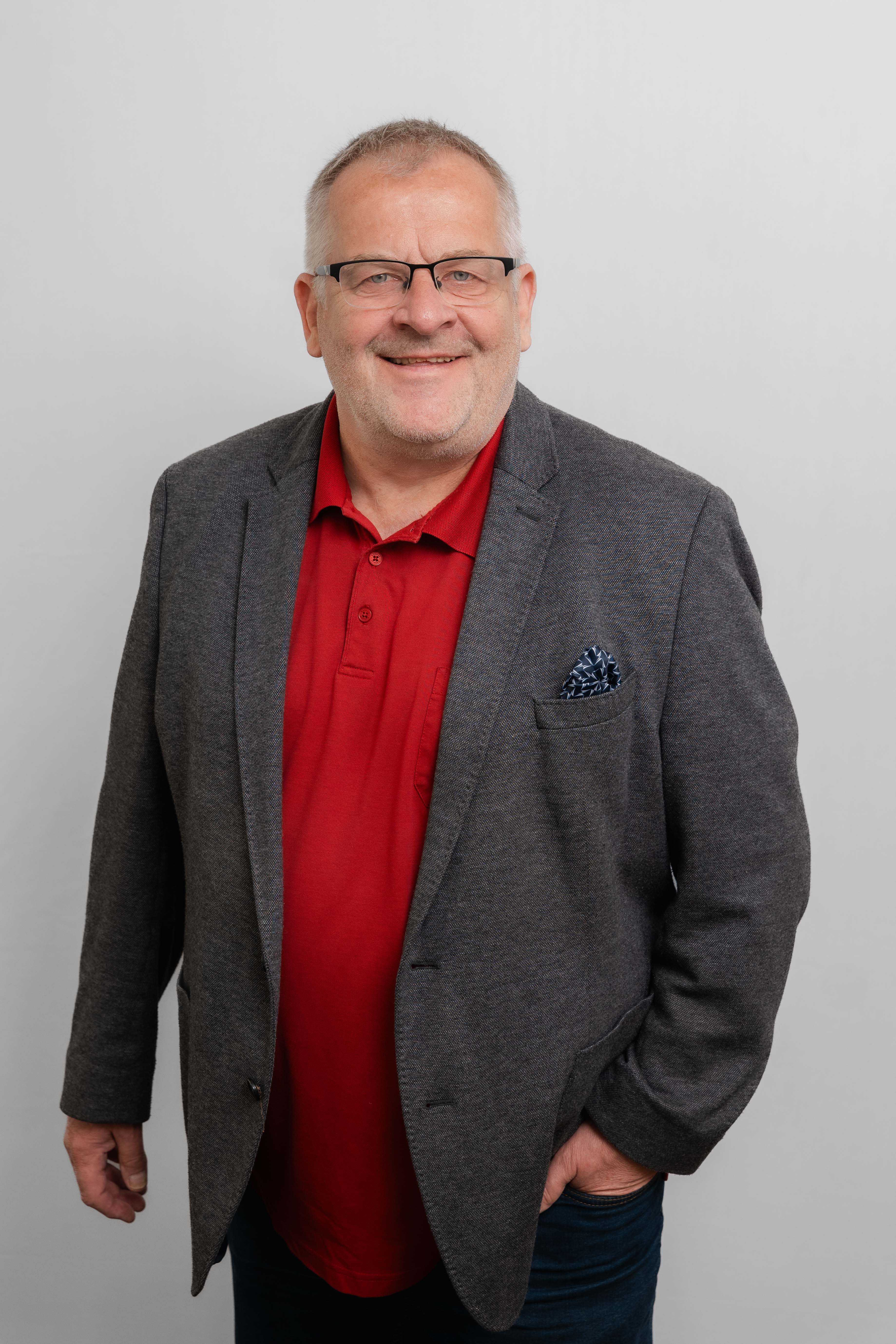Ein Mann mit Brille lacht in die Kamera. Er trägt ein rotes Poloshirt und ein dunkelgraues Sakko.