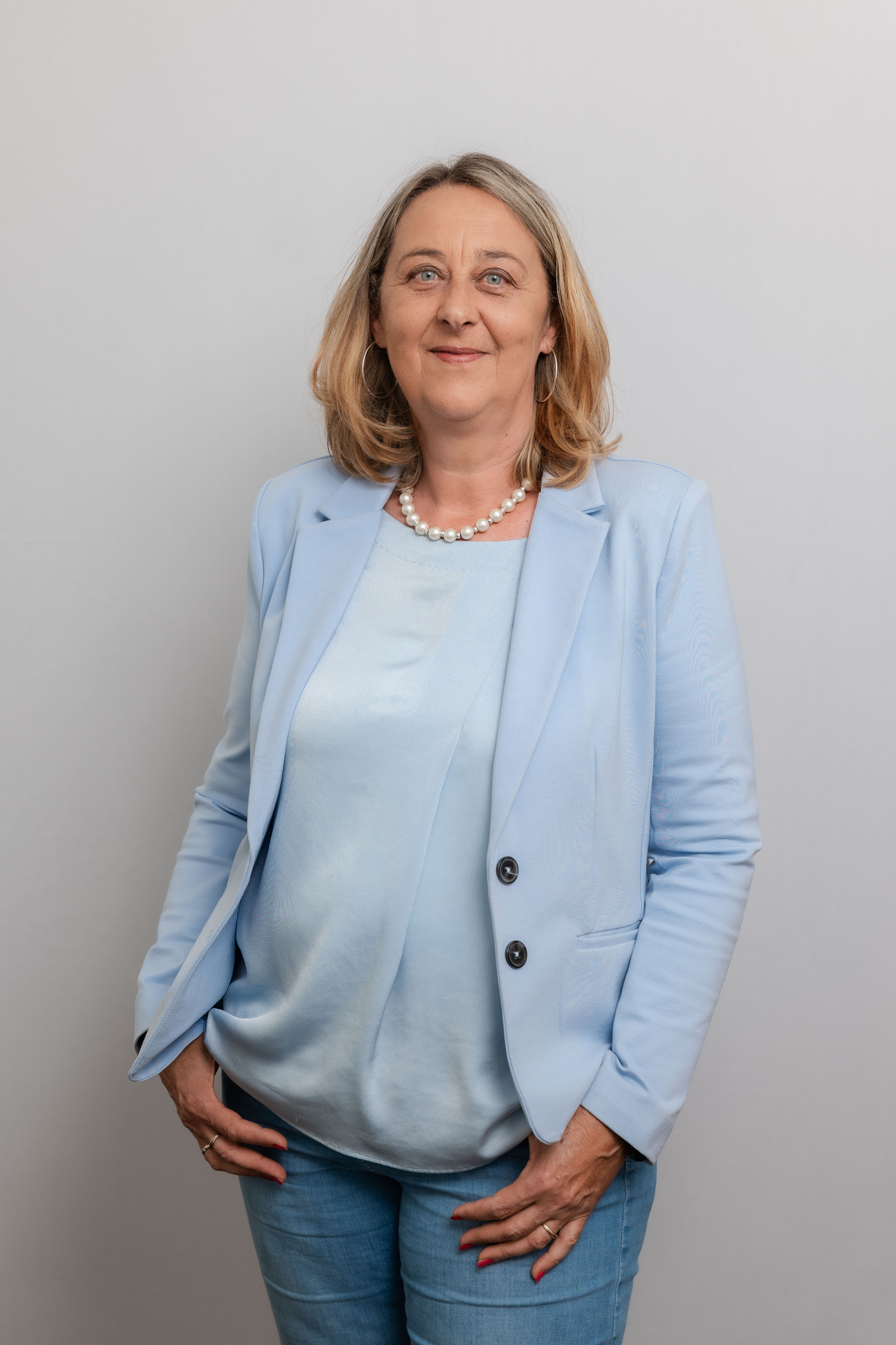 Eine Frau lacht in die Kamera. Sie trägt eine hellblaue Bluse und einen hellblauen Blazer sowie eine Perlenhalskette.