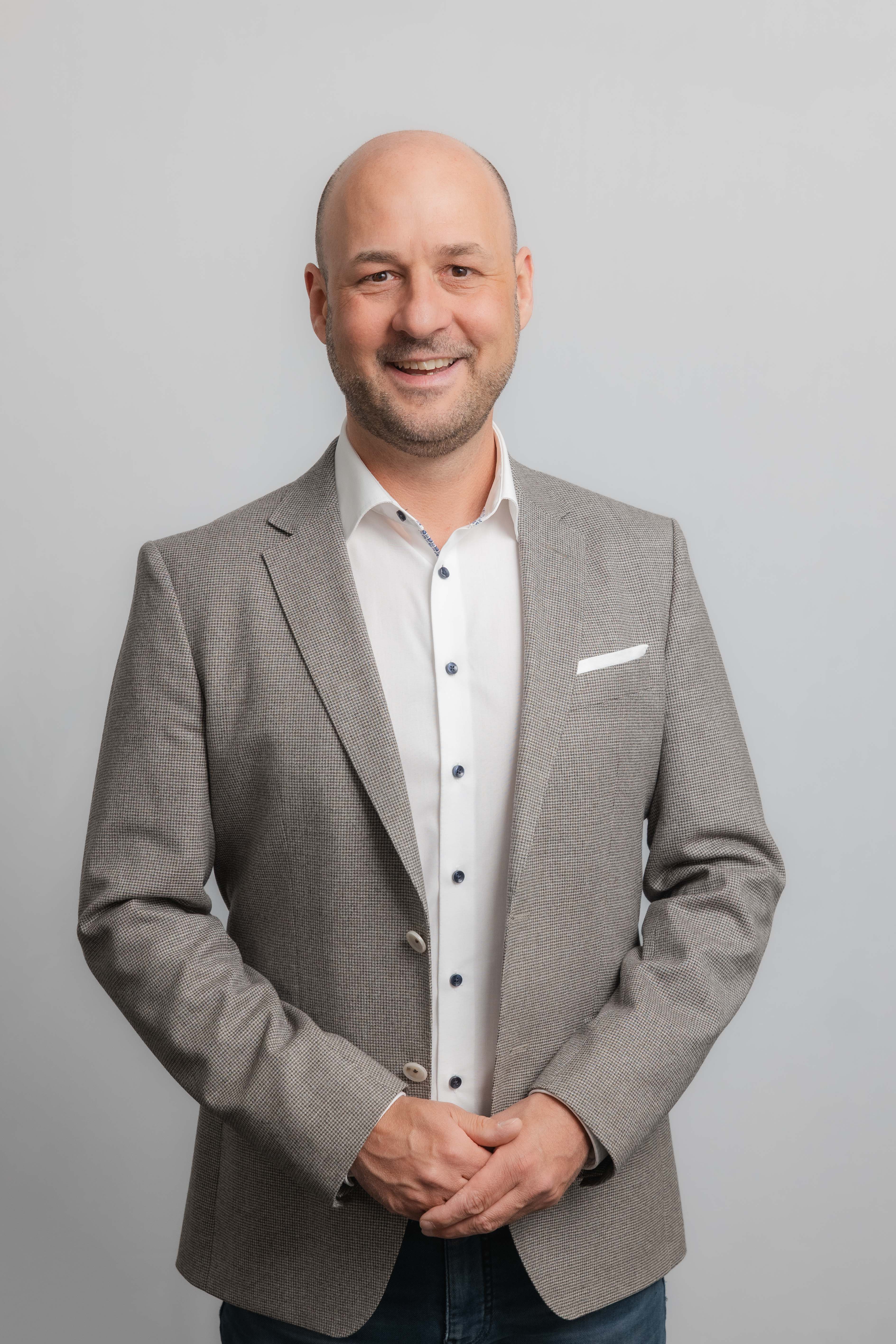 Ein Mann lacht in die Kamera. Er trägt ein weißes Hemd und ein grau-braunes Sakko.