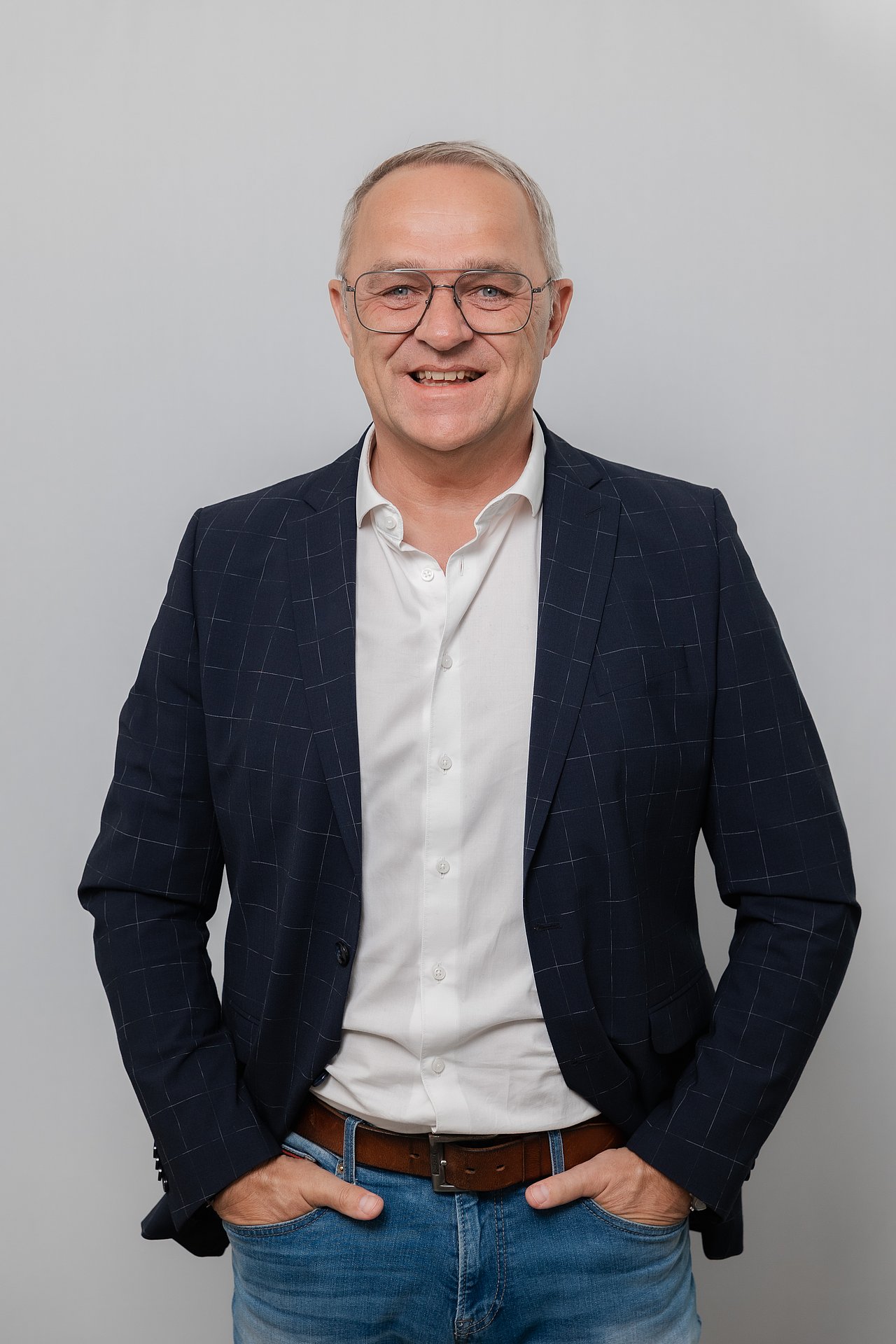 Ein Mann mit Brille lacht in die Kamera. Er trägt ein weißes Hemd mit Sakko, beide Hände stecken in den seitlichen Hosentaschen.