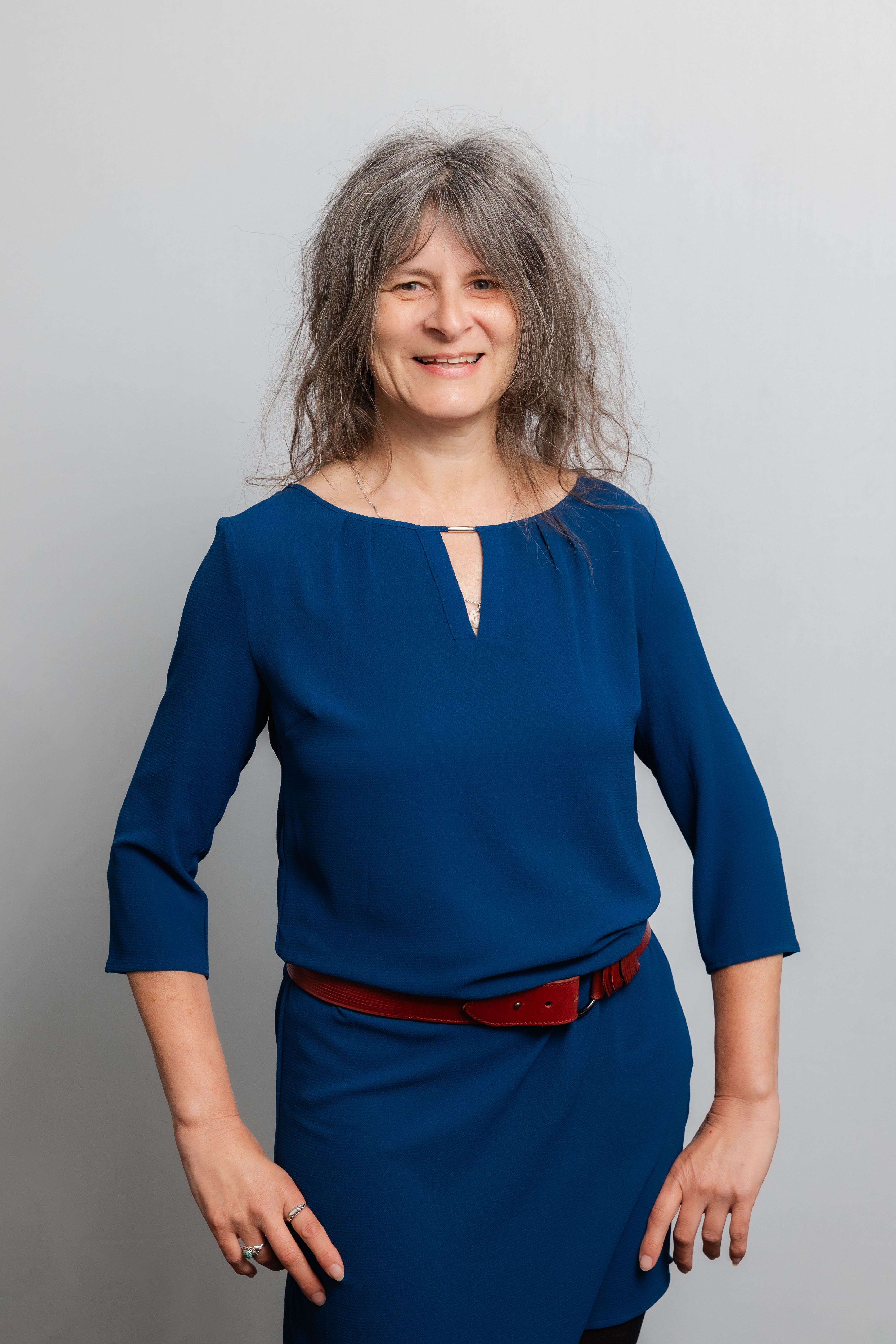 Eine Frau mit gelockten Haaren lacht in die Kamera. Sie trägt ein dunkelblaues Kleid mit einem roten Gürtel.