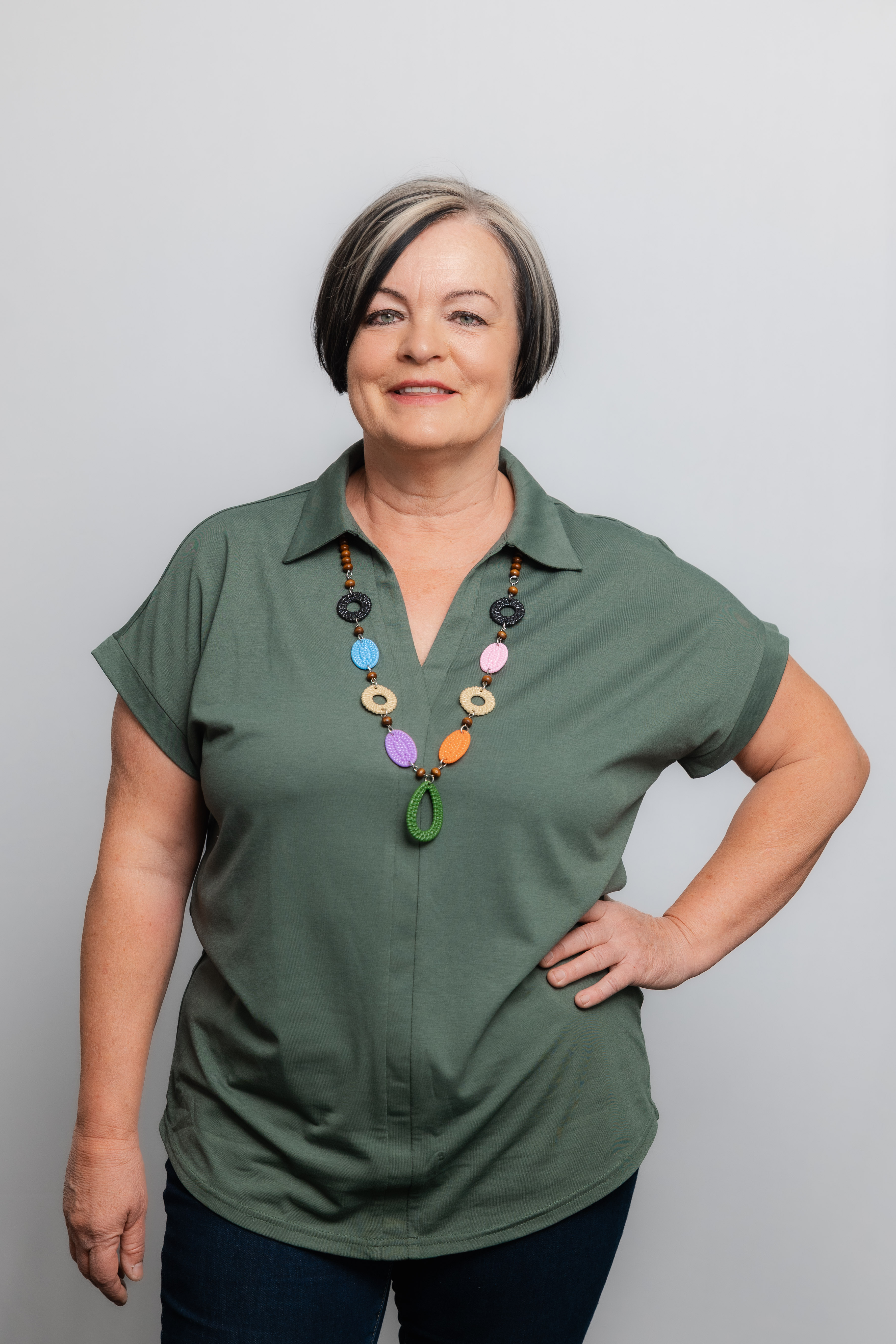 Eine Frau mit Kurzhaarschnitt lacht in die Kamera. Sie trägt eine grüne Bluse und eine bunte Halskette.