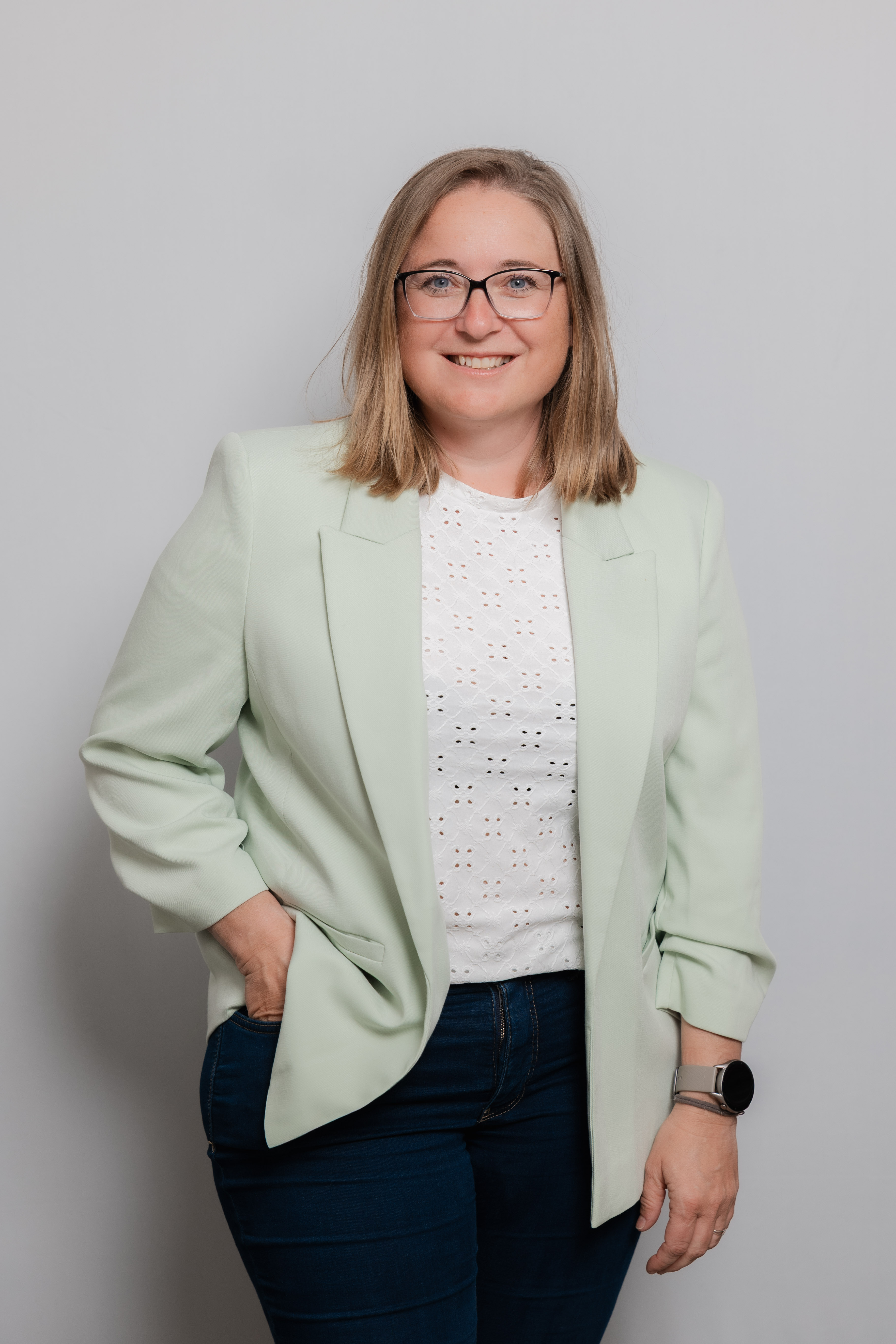Eine Frau mit Brille lacht in die Kamera. Sie trägt ein weißes Oberteil und einen mintgrünen Blazer.