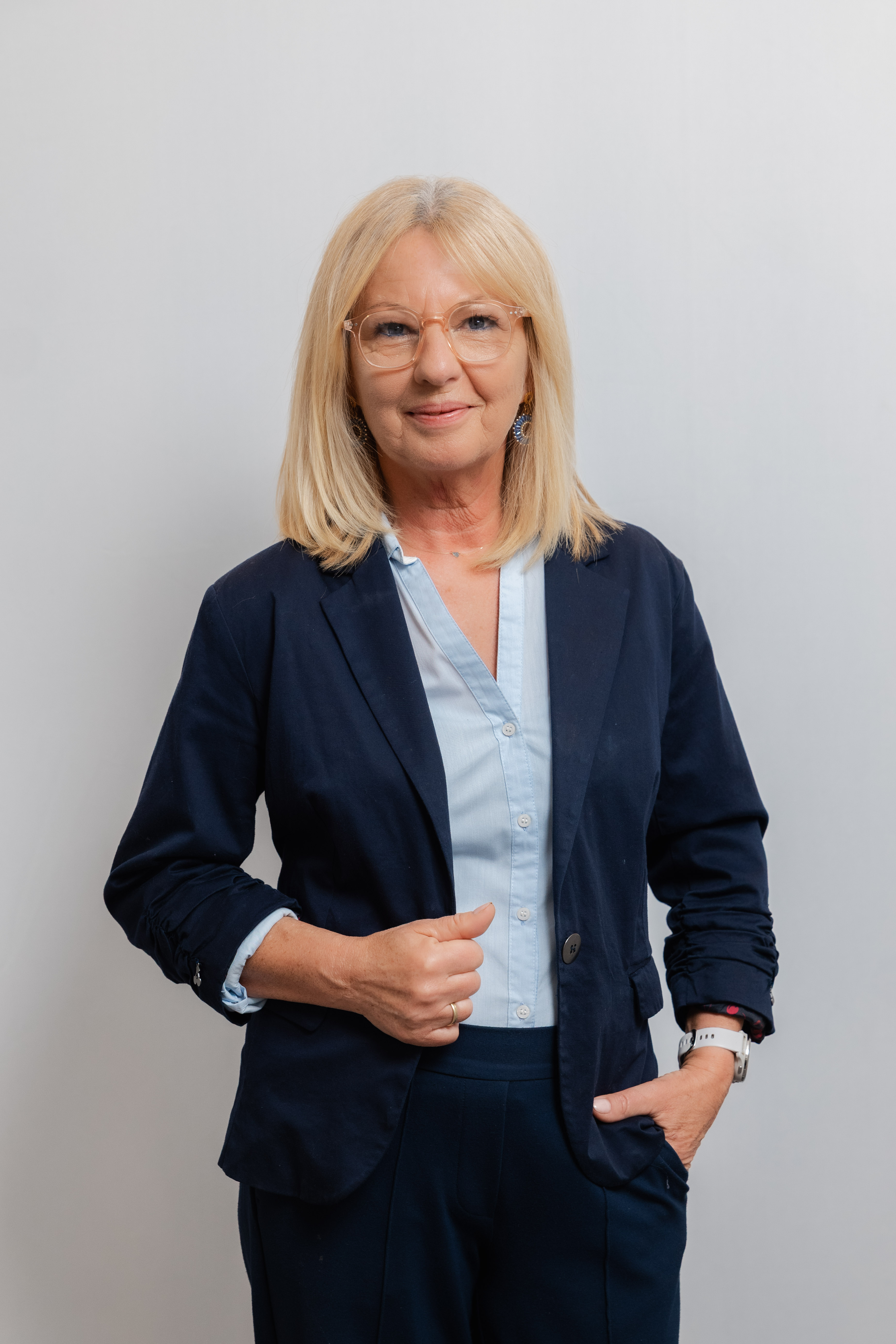 Eine Frau mit Brille lacht in die Kamera. Sie trägt einen dunkelblauen Hosenanzug und eine hellblaue Bluse.