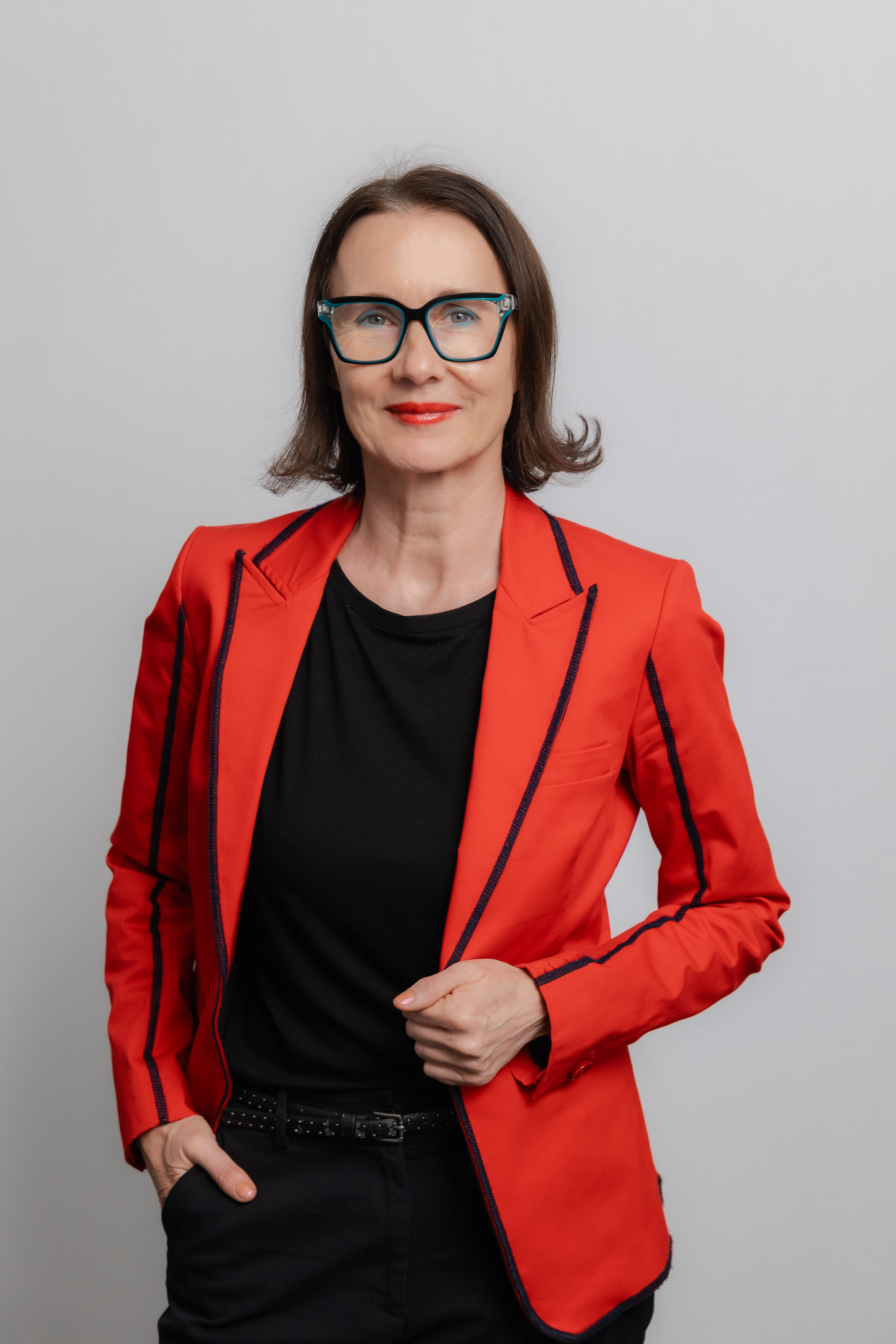 Eine Frau mit Brille lacht in die Kamera. Sie trägt ein schwarzes Oberteil und einen roten Blazer.