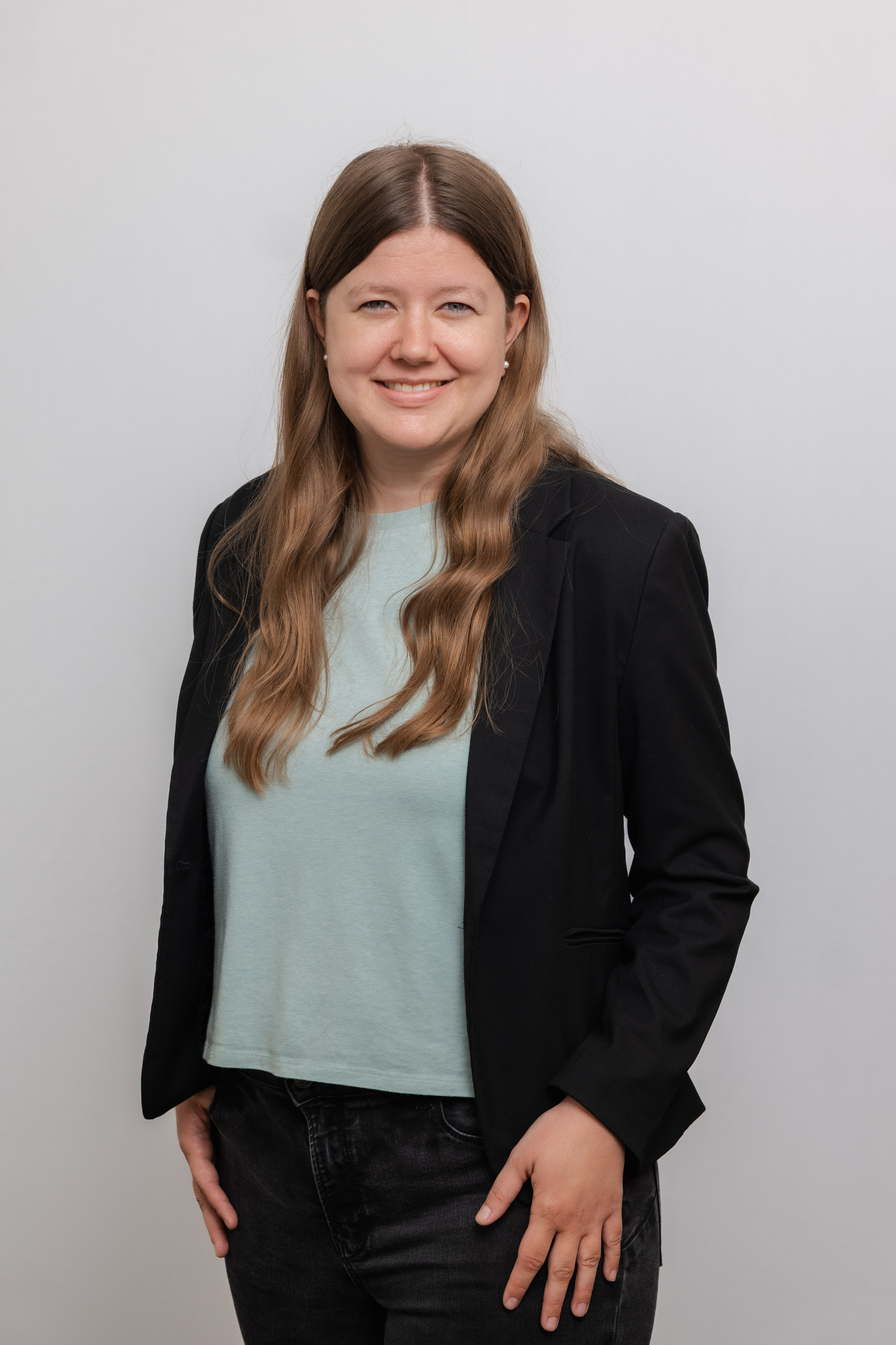 Eine Frau mit langen Locken lacht in die Kamera. Sie trägt ein hellblaues Shirt und einen schwarzen Blazer.
