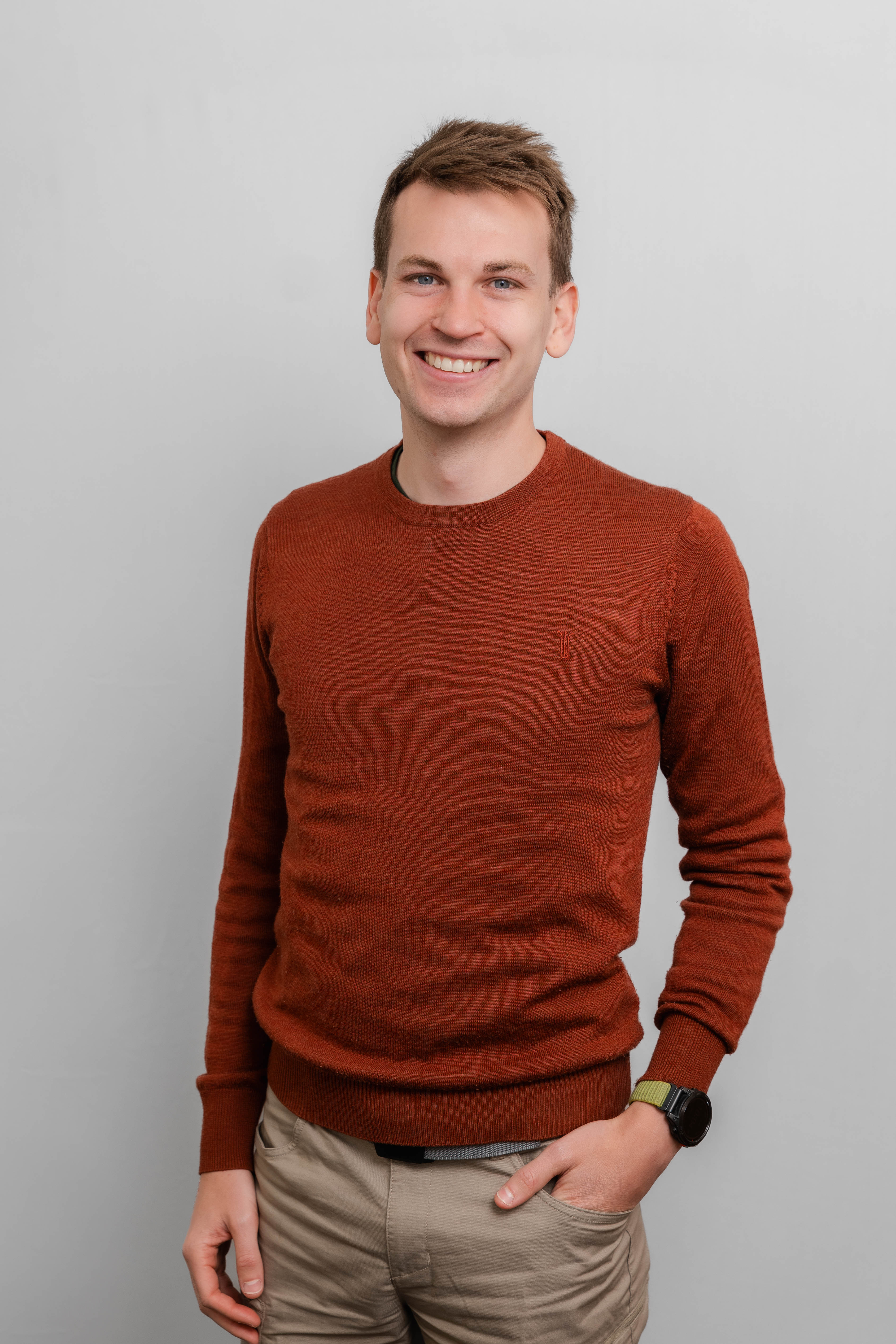 Ein Mann lacht in die Kamera. Er trägt einen roten Pullover und eine beige Hose.