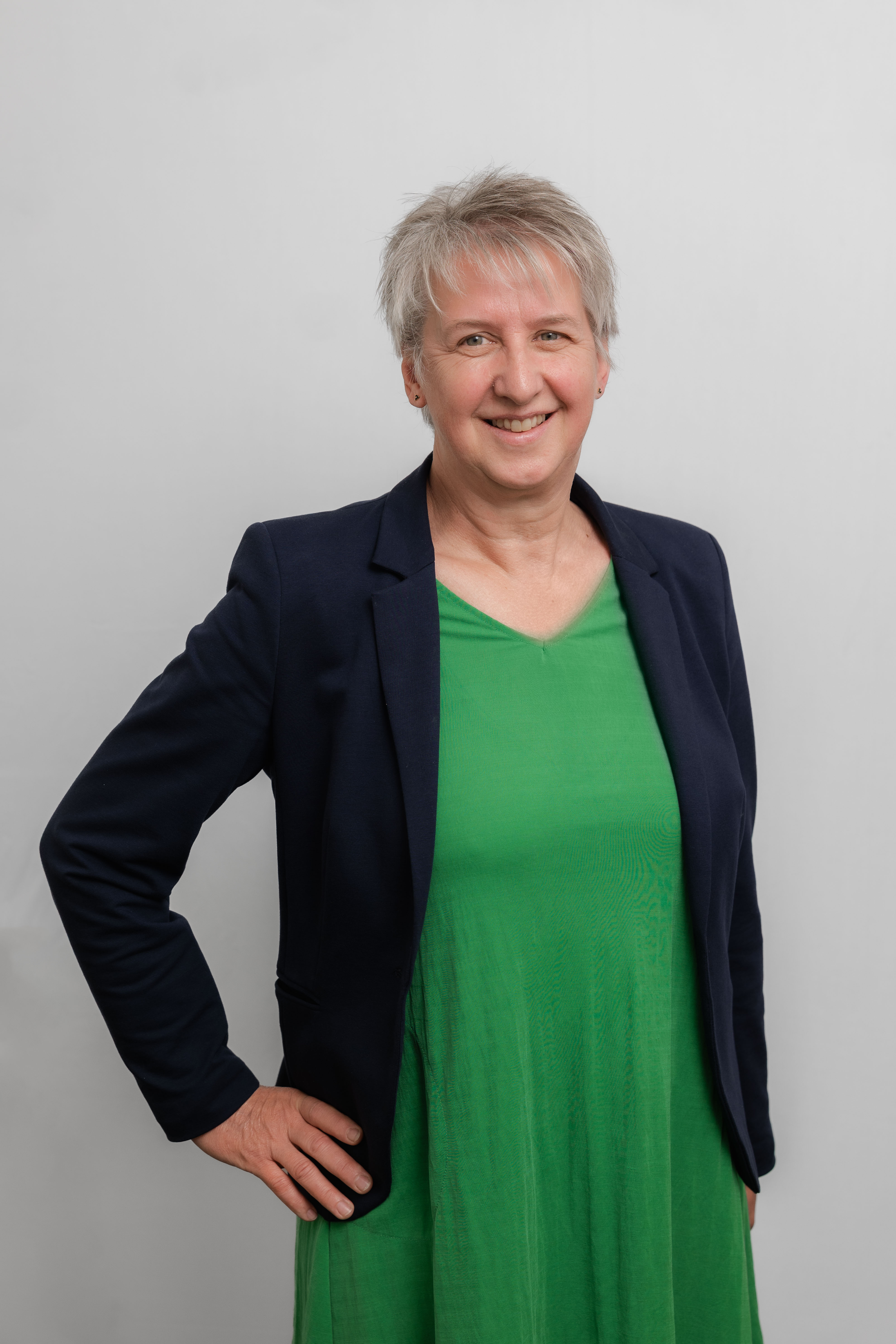 Eine Frau mit Kurzhaarschnitt lacht in die Kamera. Sie trägt ein grünes Kleid und einen schwarzen Blazer.