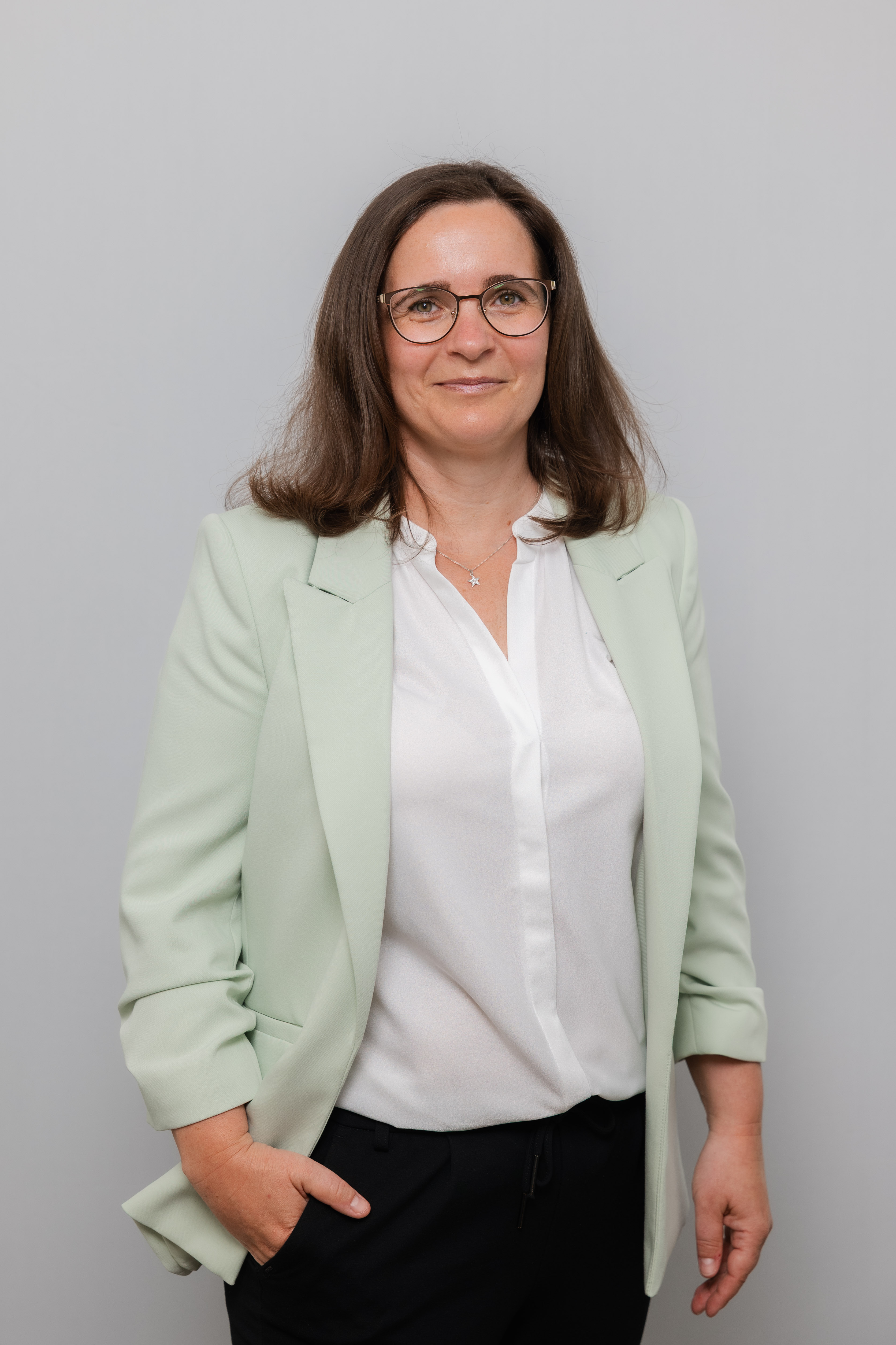 Eine Frau mit Brille lacht in die Kamera. Sie trägt eine weiße Bluse und einen mintgrünen Blazer.