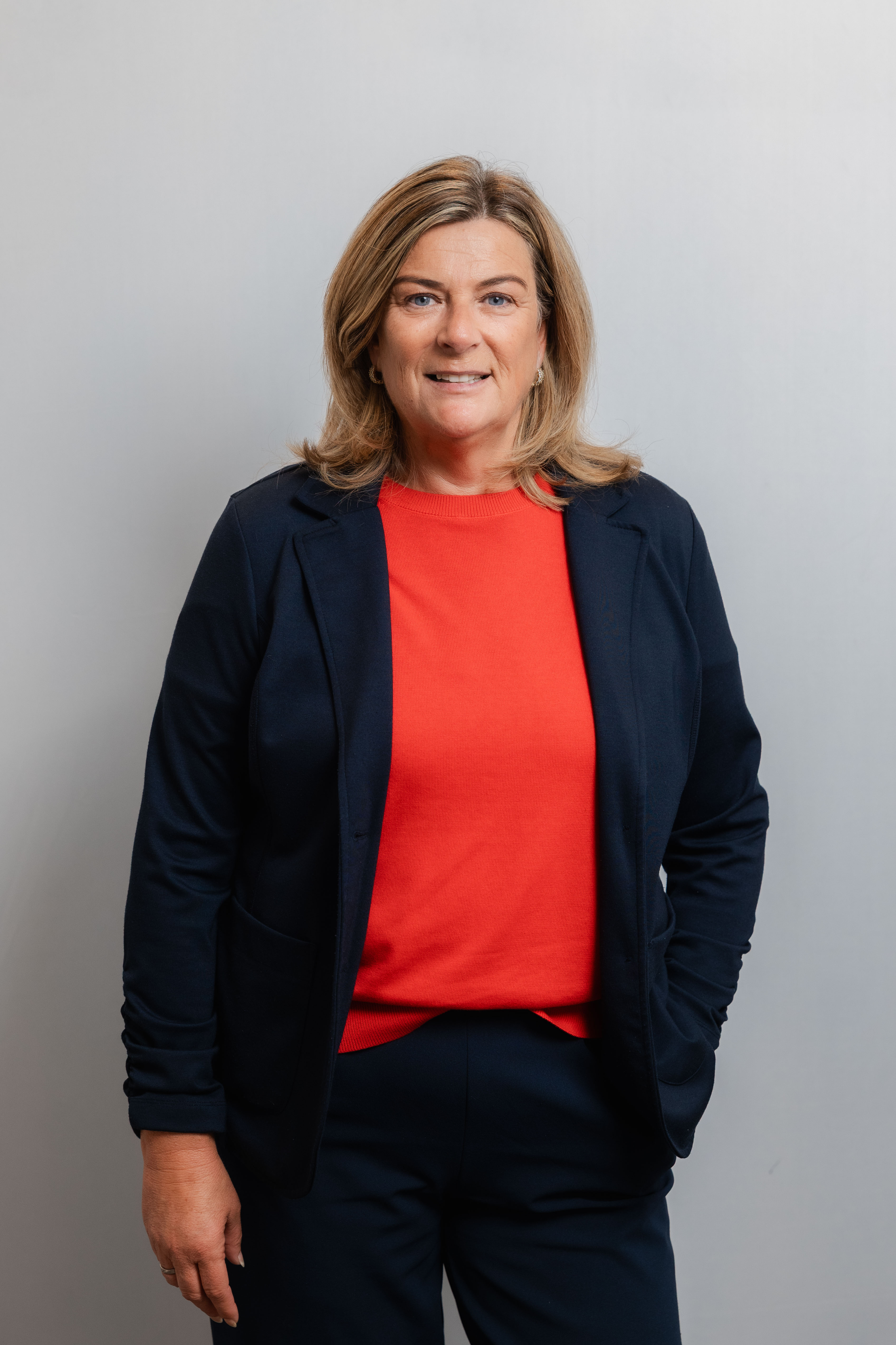 Eine Frau lacht in die Kamera. Sie trägt einen dunkelblauen Hosenanzug und ein rotes Shirt.