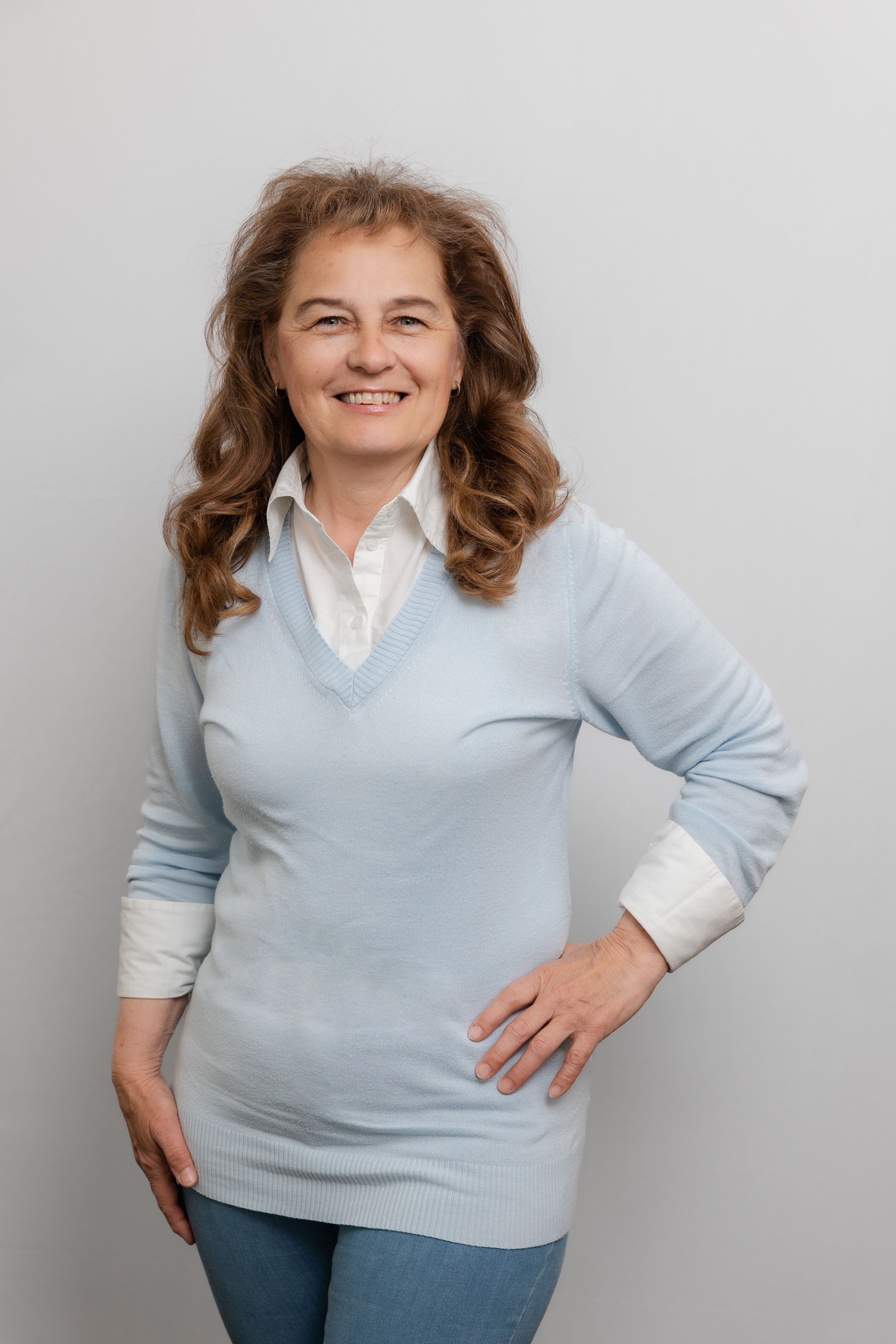 Eine Frau mit langen Locken lacht in die Kamera. Sie trägt einen hellblauen Pullover mit einer weißen Bluse darunter.