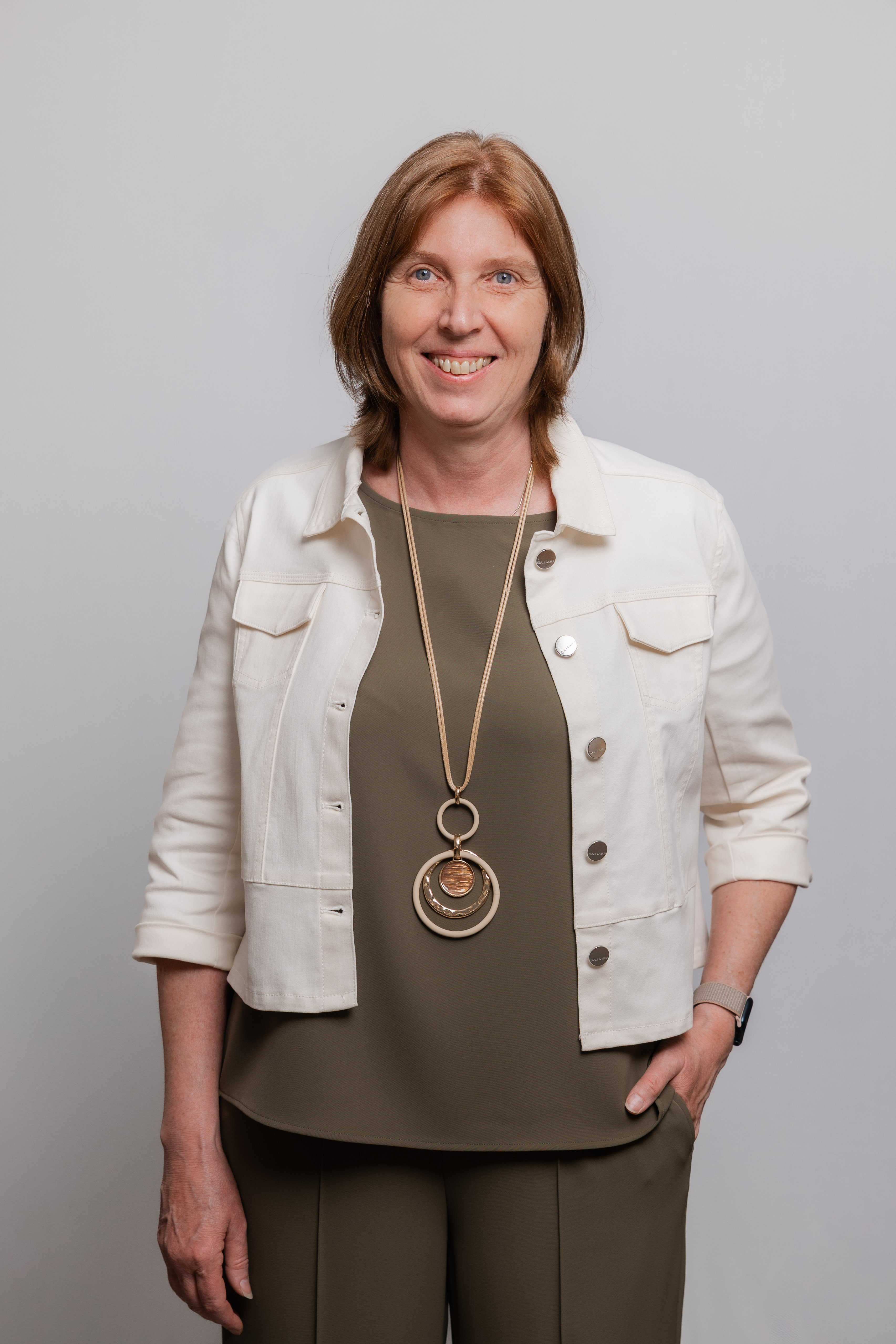 Eine Frau mit einer langen Kette um den Hals lacht in die Kamera. Sie trägt einen olivgrünen Zweiteiler und eine weiße Jacke darüber.