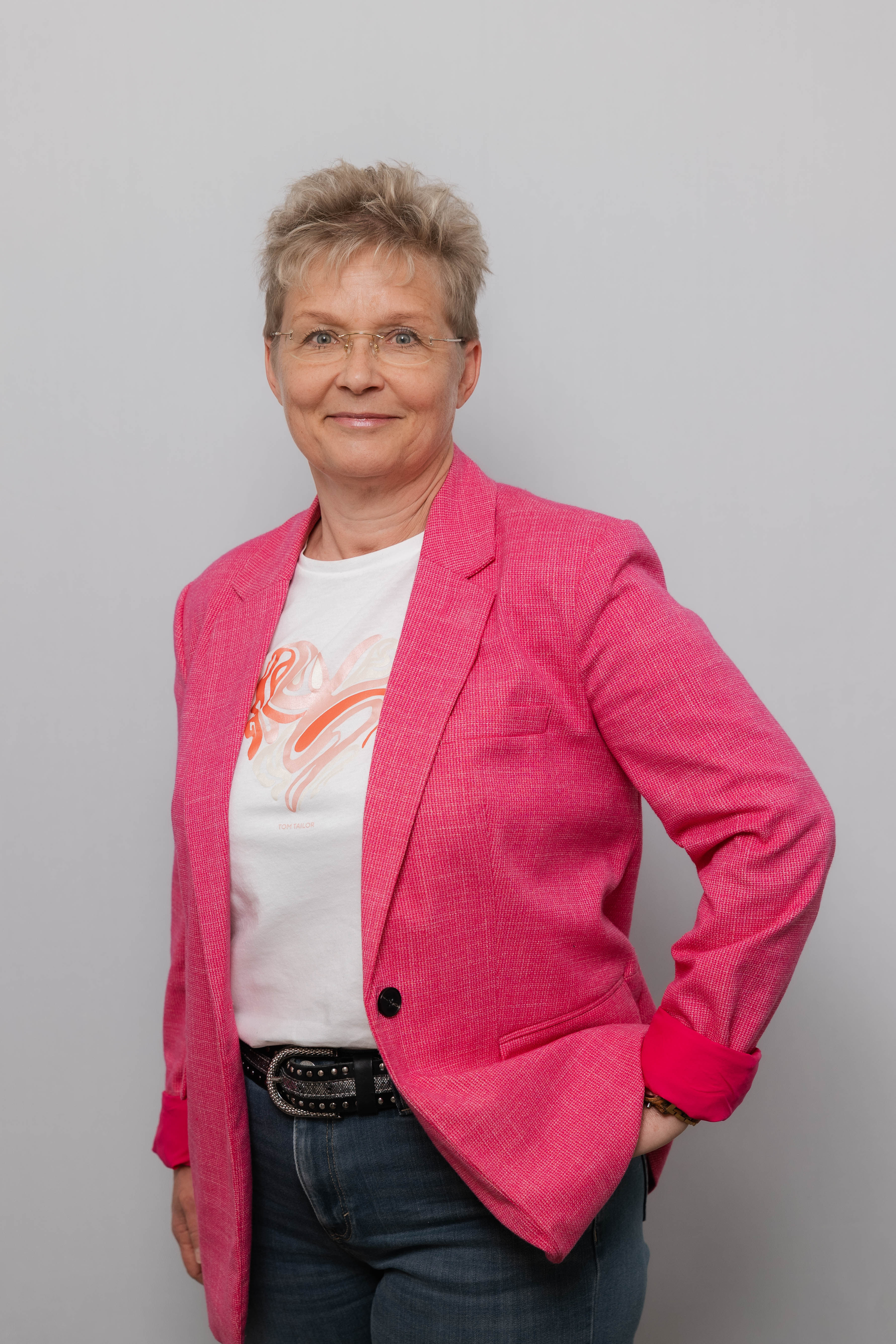 Eine Frau mit Kurzhaarschnitt und Brille lacht in die Kamera. Sie trägt einen pinken Blazer und hält eine Hand in die Hüfte gestützt.